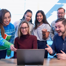 Group of excited men and women standing and sitting while staring at a laptop.