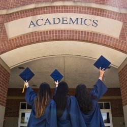 Three female students holding their graduation cap towards the academics building.